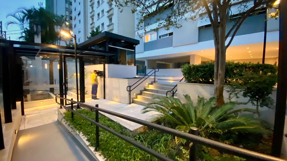 Rampa e escada de acesso iluminadas ao entardecer, com guarda-corpo preto e jardim de palmeiras.