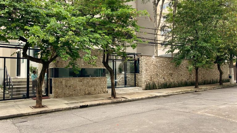 Fachada de edifício residencial com muro revestido em pedra, gradil leve e árvores na calçada.
