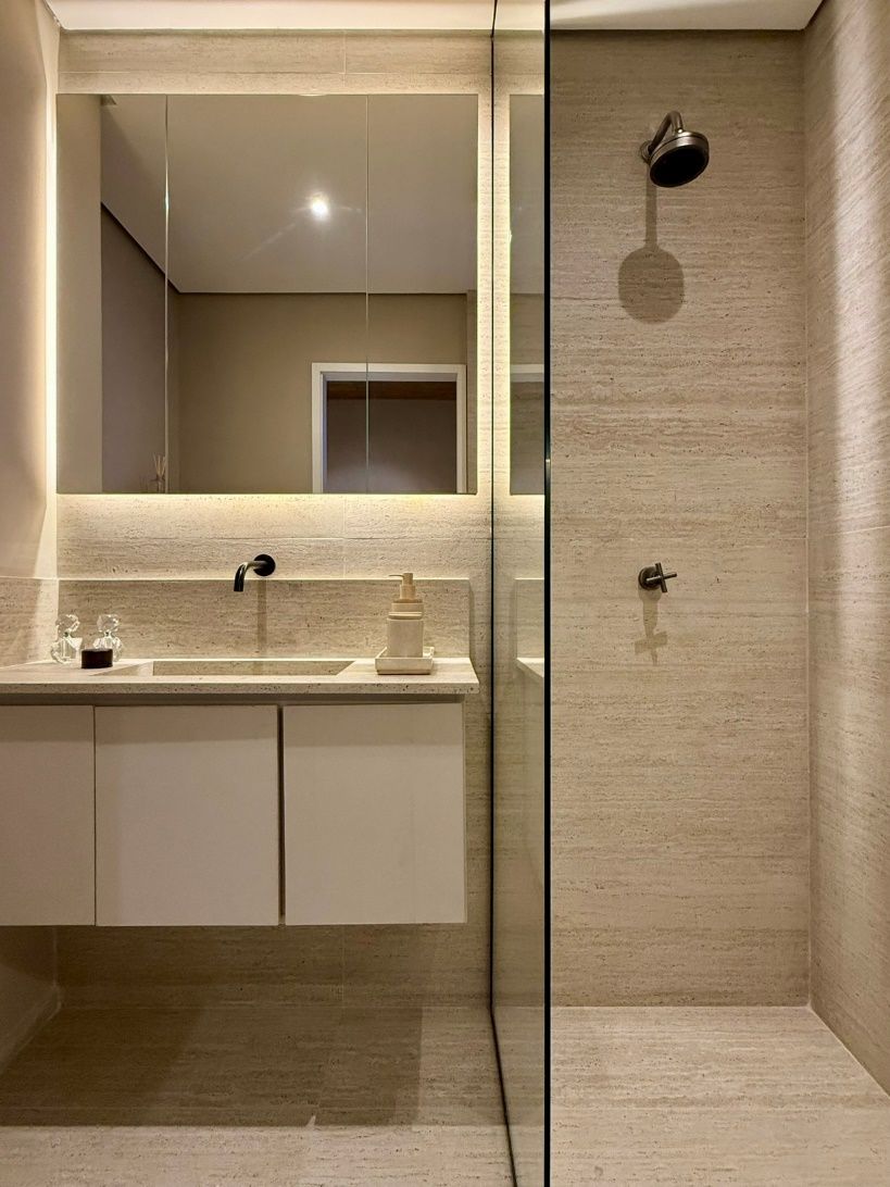Banheiro de apartamento decorado com bancada clara, espelho iluminado e box em vidro.